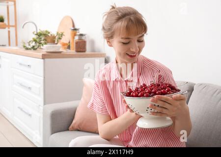 Schöne glückliche junge Frau mit Sieb voller reifer Kirschen, die auf dem Sofa in der Küche sitzt Stockfoto