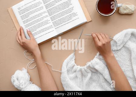 Weibliche Hände mit Stricknadeln, Buch und Tasse Tee auf beigefarbenem Hintergrund Stockfoto