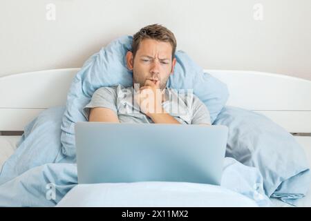 Junger, nachdenklicher Mann, der im Bett liegt und an einem Laptop arbeitet. Ernsthafter kaukasischer männlicher Freelancer, der im Internet surft und online arbeitet Stockfoto