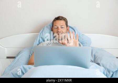 Junger fröhlicher Mann kommuniziert per Videoanruf mit Kollegen zeigt Grußgeste, während er im Bett liegt und zu Hause an einem Laptop arbeitet. Glücklich Stockfoto