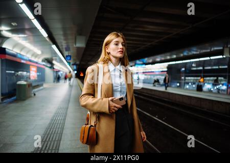 Frau, die an U-Bahn-Bahnsteig mit Handy steht, soziale Medien nutzt und Zug wartet Stockfoto