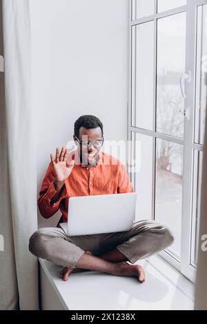 Lächelnder afroamerikanischer Mann Video, der auf Laptop durch Fenster ruft. Ein fröhlicher Mann winkt zur Webcam und nimmt an einem Videoanruf auf seinem Laptop Teil, während natürliches Licht aus dem Fenster neben ihm hereinströmt Stockfoto