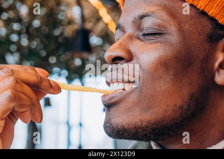 Nahaufnahme eines lächelnden afroamerikanischen Mannes, der Pommes Frites in gemütlicher Umgebung genießt. Ein lächelnder Mann, der ein französisches Frittierchen genießt, die Freude am einfachen Essen vor einem hellen Bokeh-Hintergrund Stockfoto