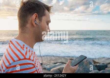 Seitenansicht eines kaukasischen jungen Mannes, der Ohrhörer an der Küste trägt und Musik hört. Hübscher Mann draußen mit Kopfhörern am Strand Stockfoto