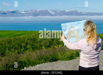 Junge Touristin schaut sich die Karte der isländischen Westfjorde in idyllischer Natur mit schneebedeckten Bergen im Hintergrund und Fjord an. Stockfoto