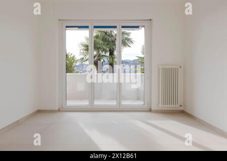 Leeres Zimmer mit einem großen Fenster im Hintergrund, das auf den Balkon führt. Rechts an der weißen Wand ist ein Heizkörper oder eine Heizung zu sehen. Stockfoto