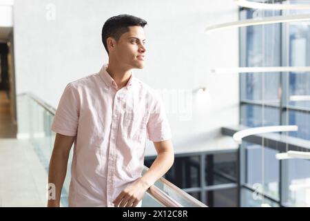 Ein junger, birassischer Mann, der sich auf einem Geländer stützt und in einem modernen Geschäftsbüro mit Kopierraum zur Seite blickt Stockfoto