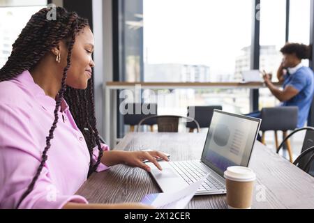 Eine junge Frau mit geflochtenen Haaren arbeitet in einem modernen Geschäftsbüro an einem Laptop Stockfoto