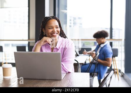 Junge Frau mit geflochtenem Haar, die am Laptop arbeitet, lächelnd, in einem modernen Geschäftsbüro Stockfoto