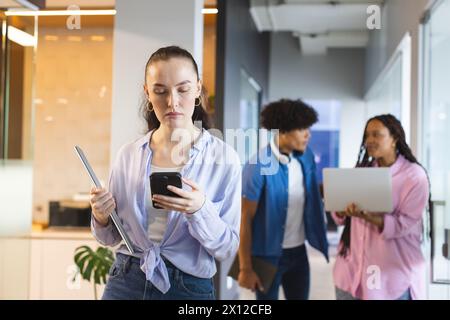 Kaukasische Frau, die ihr Smartphone überprüft, birassische Kollegen, die in einem modernen Geschäftsbüro laufen Stockfoto