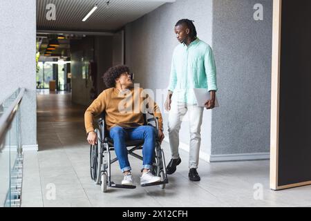 Afroamerikanischer Mann, der neben birassischem Mann im Rollstuhl in einem modernen Geschäftsbüro läuft Stockfoto