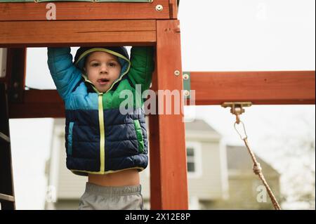 Glücklicher Junge, der im Winter draußen auf dem Spielset im Mantel schwingt Stockfoto