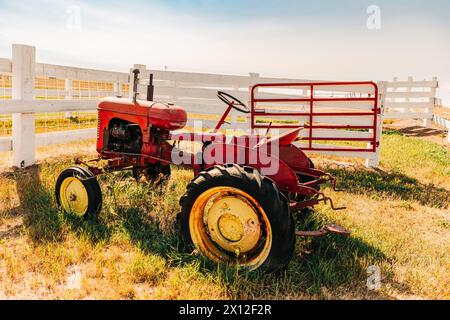 Vintage verwitterter roter Traktor in der Nähe des weißen Pfahlzauns an sonnigen Tagen Stockfoto