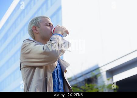 Junger blonder argentinischer lateinmann, elegant mit Jacke und Hemd, abgelenkt draußen stehend, Kaffee trinken und sich umsehen mit Gebäuden in BA Stockfoto