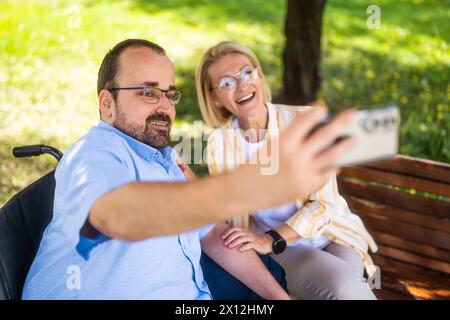 Ein Mann im Rollstuhl verbringt Zeit mit seiner Mutter im Park. Sie machen Selfie. Stockfoto