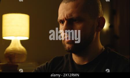 Skeptischer, verdächtiger junger Mann mit ernstem Ausdruck, der in einem dunklen Zimmer sitzt. Stockfoto