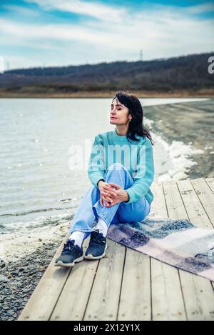 49-jährige Russin im Pullover, die auf einem Pier sitzt und mit geschlossenen Augen denkt Stockfoto