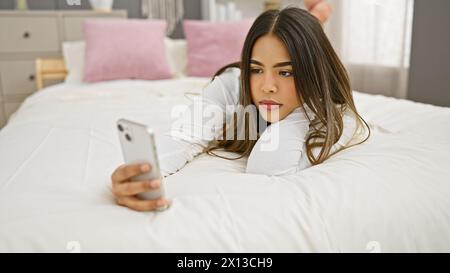 Eine junge hispanische Frau liegt im Bett und hält ein Smartphone in der Hand, um eine entspannte Indoor-Szene zu zeigen. Stockfoto