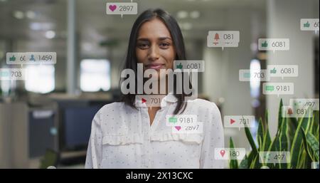 Bild von wechselnden Zahlen, Symbolen in Benachrichtigungsbalken, lächelnder birassischer Frau, die im Büro steht Stockfoto
