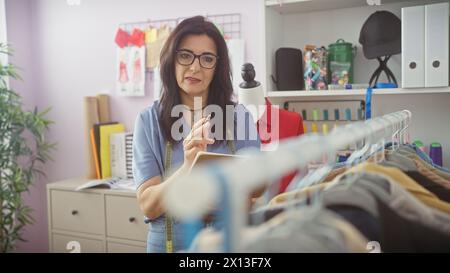 Eine Schneiderin mittleren Alters in Brille steht in ihrem Atelier mit bunten Garnen und Outfits. Stockfoto