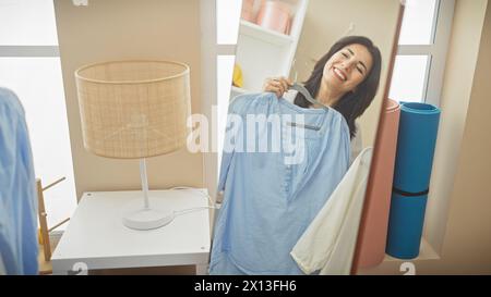 Lächelnde hispanische Frau mittleren Alters, die blaues Hemd in einem hellen Ankleidezimmer anprobiert Stockfoto