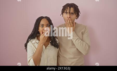 Ein überraschtes Paar mit einer Frau und einem Mann, die vor einem rosafarbenen Hintergrund stehen und ihren Mund mit Händen bedecken. Stockfoto
