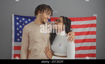 Glückliches Paar mit „ich habe gestimmt“-Aufklebern, die vor einer amerikanischen Flagge stehen und die Teilnahme an den Wahlen in den usa symbolisieren. Stockfoto