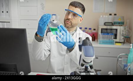 Ein junger bärtiger Mann untersucht eine Flasche mit grüner Flüssigkeit in einem Labor mit Schutzbrille und Handschuhen. Stockfoto