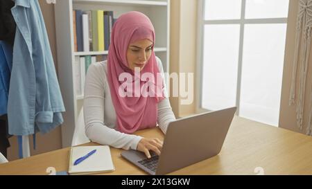 Eine fokussierte Frau im Hijab arbeitet an ihrem Laptop an einem Schreibtisch mit Büchern und einem Notizblock. Stockfoto