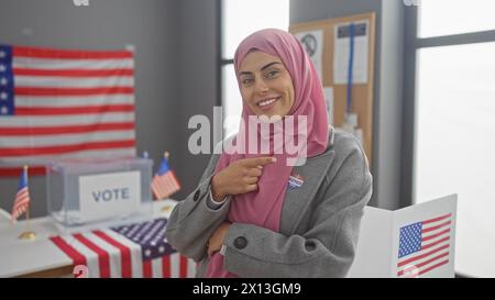 Eine selbstbewusste junge Frau, die einen Hijab trägt, zeigt auf ihren „ich habe gewählt“-Aufkleber in einem US-Wahlstudienzimmer, der mit amerikanischen Fahnen geschmückt ist. Stockfoto