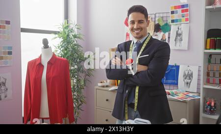 Lächelnder Mann mit Bart in Schneiderei, der Arme kreuzt, umgeben von bunten Stoffmustern und Nähgeräten. Stockfoto