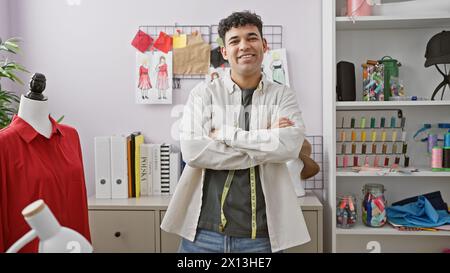 Ein lächelnder junger Mann mit überkreuzten Armen steht in einem Schneidergeschäft, umgeben von bunten Garnen, Kleidungsstücken und Nähzubehör. Stockfoto