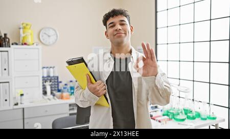 Ein hübscher Mann mit einem Klemmbrett, das in einem Labor ein „OK“-Zeichen gibt. Stockfoto