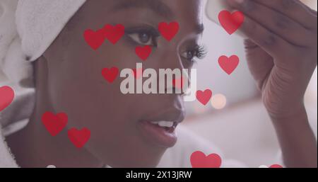 Bild von roten Herzen über glücklicher afroamerikanischer Frau, die zu Hause das Gesicht reinigt Stockfoto