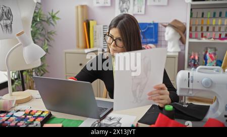 Eine fokussierte Modedesignerin arbeitet in einem farbenfrohen Atelier, umgeben von Nähaccessoires, Skizzen und einem Laptop. Stockfoto