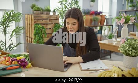 Eine junge hispanische Frau arbeitet an einem Laptop in einem Blumenladen voller bunter Tulpen und grüner Pflanzen. Stockfoto