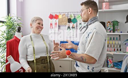 Zwei Schneider, eine Frau und ein Mann, unterhalten sich freudig in einem pulsierenden Schneidergeschäft voller Mode-Design-Skizzen und Nähausrüstung. Stockfoto