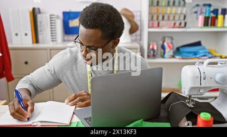 afroamerikanischer Schneidermann macht Notizen in einem modernen Atelier mit Nähmaterial und Laptop Stockfoto