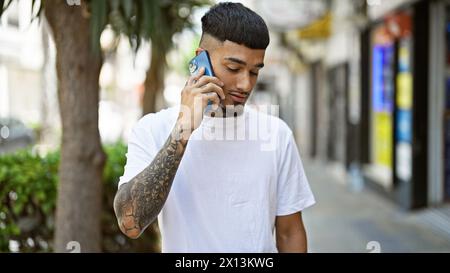 Cooler, tätowierter junger latino-Mann, der ernsthaft in Telefongespräche vertieft ist, draußen auf der sonnendurchfluteten Stadtstraße steht und entspannte Vibes ausstrahlt, während er s verwendet Stockfoto