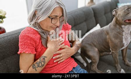 Die herzerstreute grauhaarige Frau mittleren Alters leidet in ihrem gemütlichen Wohnzimmer unter einem Angriff, während sie mit einem geschätzten Hund auf einem bequemen Sofa sitzt. Stockfoto