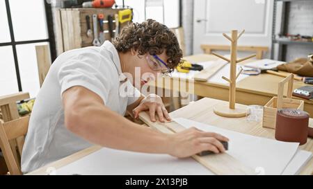 Ein fokussierter junger hispanischer Mann mit lockigen Haaren arbeitet in einer Innenwerkstatt an der Tischlerei. Stockfoto
