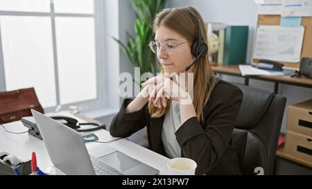 Eine professionelle junge Frau, die ein Headset trägt, konzentriert sich, während sie in einem modernen Büro an ihrem Laptop arbeitet. Stockfoto