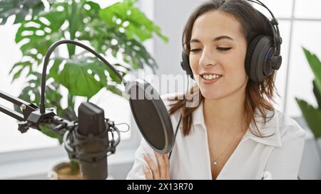 Die lächelnde hispanische Frau mit Kopfhörern nimmt Podcast in hellem Studio auf Stockfoto