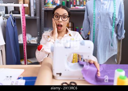 Junge arabische Schneiderin, die im Atelier arbeitet, ängstlich und schockiert, überrascht und verblüfft mit Händen im Gesicht Stockfoto