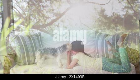 Zusammengesetztes Bild von hohen Bäumen im Wald gegen das kaukasische Mädchen, das mit ihrem Hund auf der Couch spielt Stockfoto