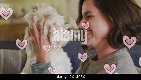 Mehrere rosafarbene Herzsymbole schwebten gegen eine weiße Frau, die ihren Hund zu Hause küsst Stockfoto