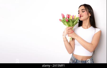 Junge Frau in lässigem Outfit hält einen Strauß rosa Tulpen und riecht sie auf weißem isoliertem Hintergrund Stockfoto