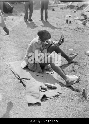 VICTORY MARCHERS CAMP IN LONDON: UNTERKUNFT IN KENSINGTON GARDENS, LONDON, ENGLAND, UK, 1946 - Ein westafrikanischer Soldat rasiert sich in Vorbereitung auf die SIEGESPARADE in London. Er sitzt auf einer Decke auf dem Gras neben dem Zeltlager, in dem er und viele andere Soldaten in Kensington Gardens untergebracht sind Stockfoto