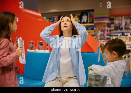 Müde Mutter hält den Kopf, während Kinder nach mehr Spielzeug fragen Stockfoto