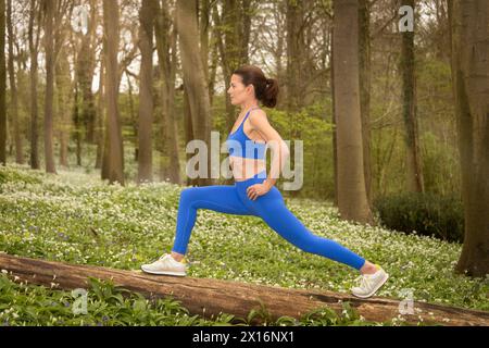 Sportliche Frau, die Dehnungsübungen auf einem Baumstamm in einem Wald macht, wilder Knoblauch und Blumen Hintergrund. Fitness-Konzept für Frühling und Sommer. Stockfoto